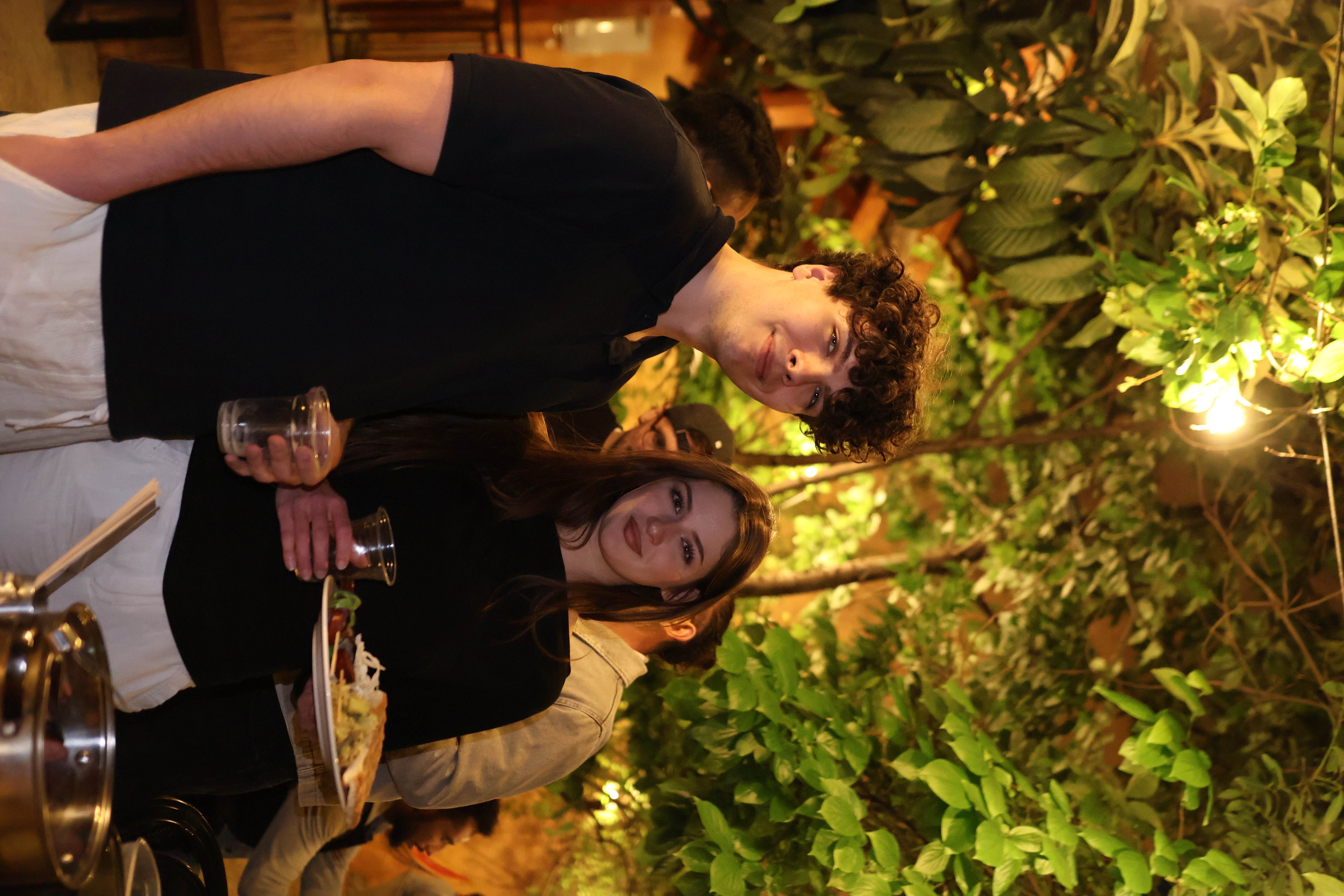 A young man and woman holding drinks and food while posing outdoors.
